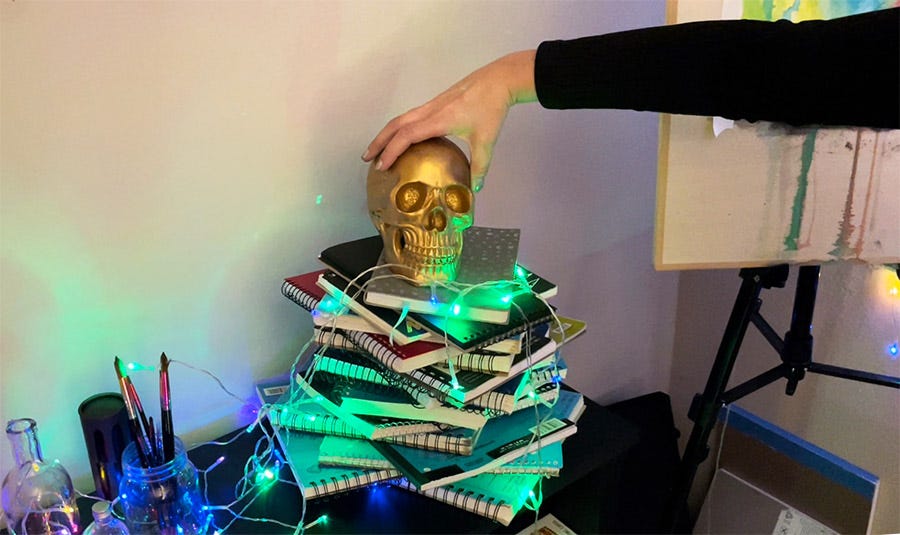 My hand placing a gold skull onto a pile of sketchbooks shaped like a Christmas tree with green lights.