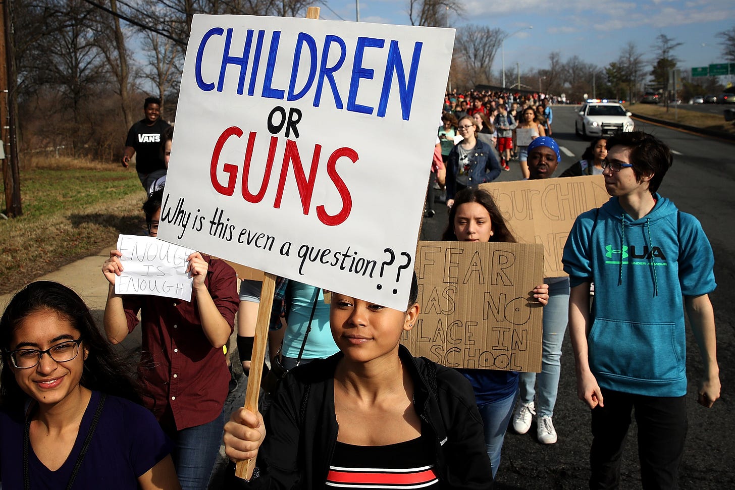 A group of teenagers protesting outdoors against gun violenve after the Santa Fe High School shooting. The dark-skinned girl in the front of the procession is holding a sign that says "CHILDREN OR GUNS Why is this even a question??" Other visible signs say "Enough is Enough" and "Fear Has No Place In School."