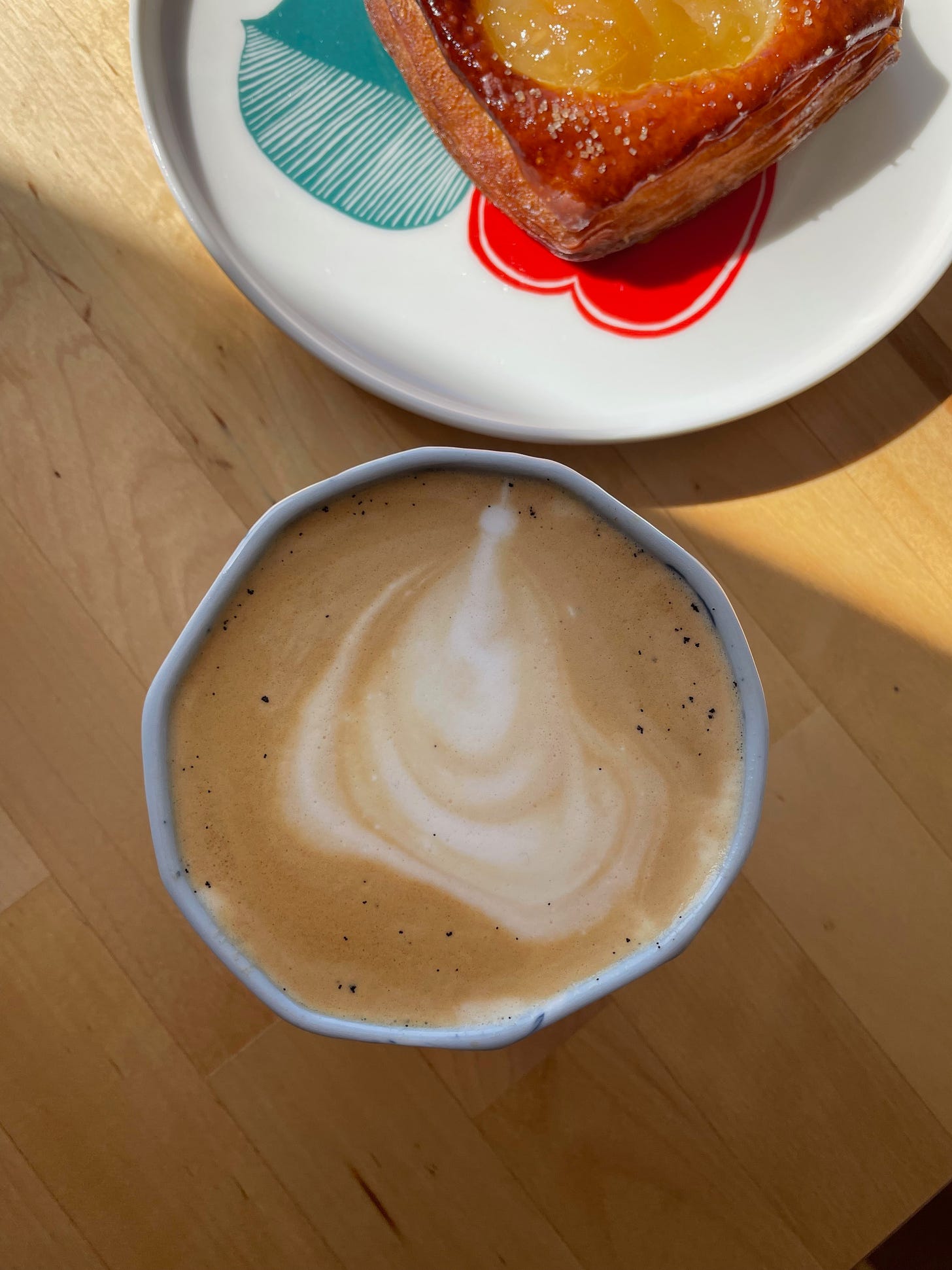 Close up of a latte with speckles from a vanilla bean syrup. A pear pastry in the sun in the background.