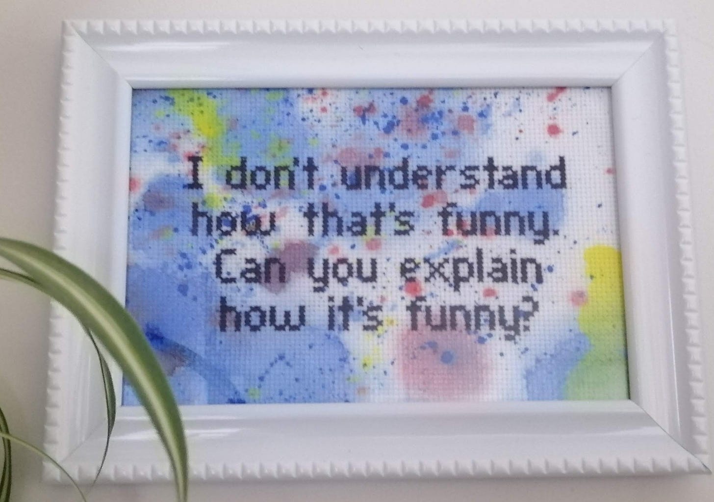 Completed cross stitch in a frame on a splotchy rainbow fabric. The text reads "I don't understand how that's funny. Can you explain how it's funny?"