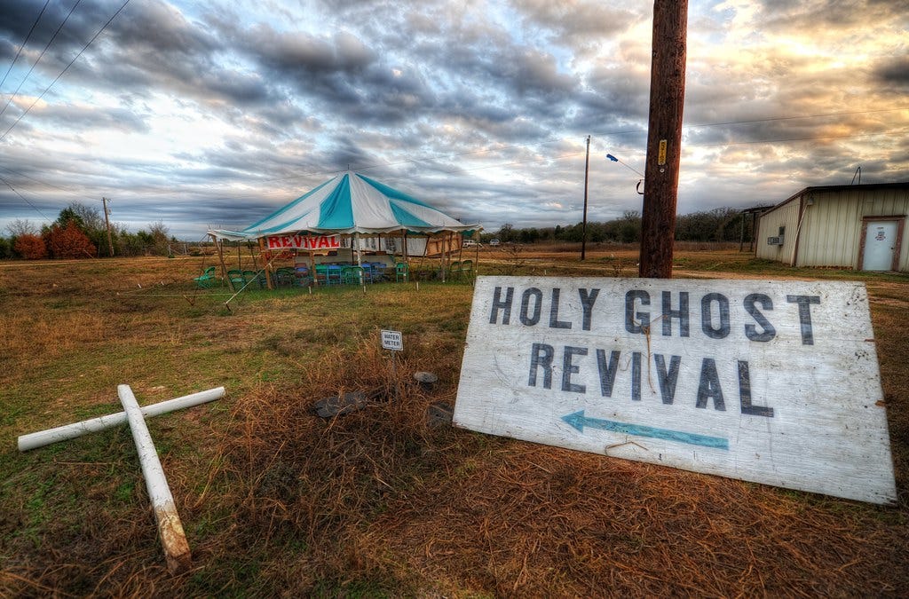 A distressed signed painted white reads "HOLY GHOST REVIVAL" while a white PVC pipe cross lies on the ground next to t The ground is damp with overgrown and sometimes dying weeds. In the background there is a small storage facility and a white and blue striped tent nearby, with chairs and a red "revival" sign present. The sky is during the sunset is so gorgeous.