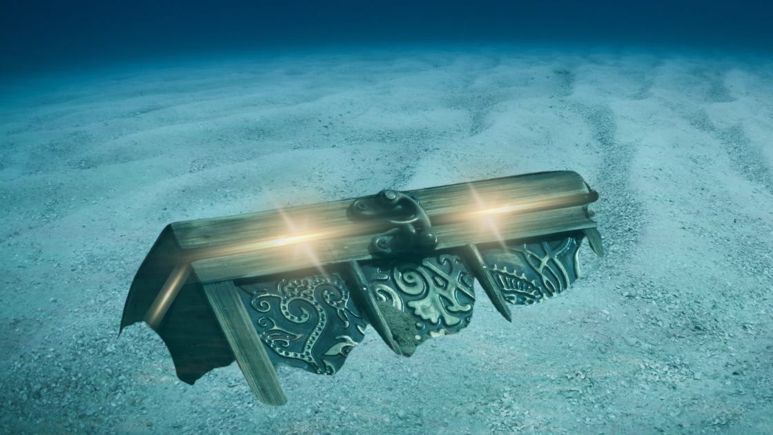 An ornate treasure chest lies half-buried in the sand of the sea floor. From the cracks of the locked lid, beams of golden light peek through.