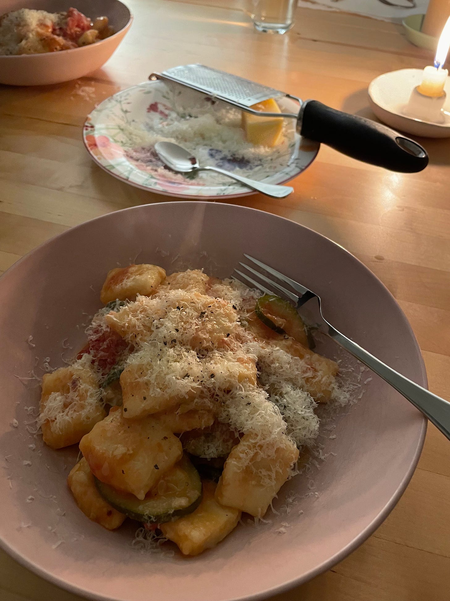 Bowl of potato gnocchi topped with parmesan cheese. It's in a tomato and zucchini sauce. Extra cheese on a plate in the background with a microplane.