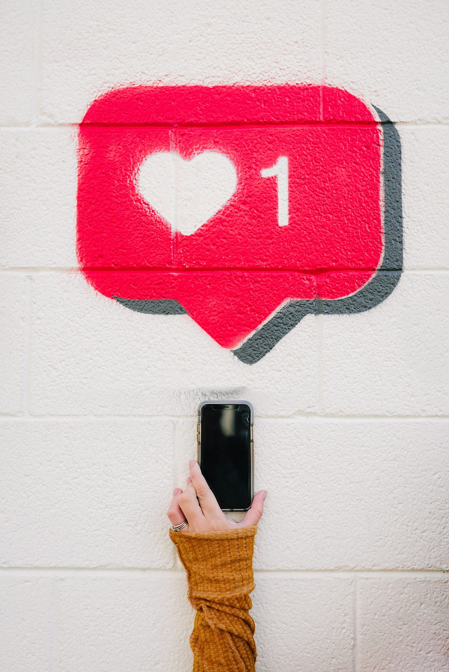 A person's hand holding a smartphone in front of a white brick wall with a red social media 'like' icon painted on it, showing a white heart and the number '1'.