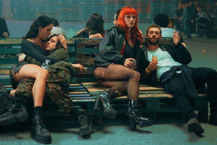 In an emerald-green hued warehouse, piles of party-goers sit on wood palettes, four of whom face forward. They are all in black and grey and neutrals, bodies entwined, except for one of them, who is beautiful and stunning with long bright-red hair.