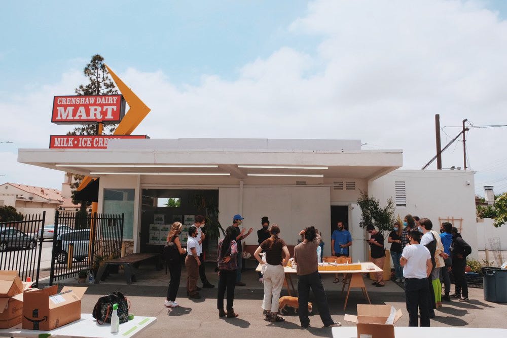 A small market with a big vintage sign that reads "Crenshaw Dairy Mart" The building has been converted into a community art gallery and people are standing around outside talking and eating