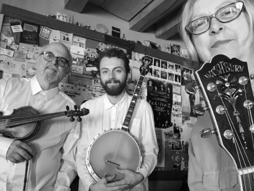 A fiddler, banjoist and guitar player, two men, one woman, smiling at the camera.