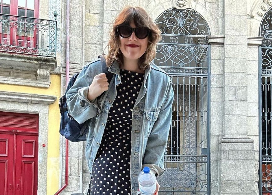 photo of Jamie Feldman, the guest from TTFA Episode "Jamie The Good Times Gal." Jamie is wearing a bold burgundy lipstick and big black sunglasses. Her brown hair is cut into a shag haircut, and she's wearing a black and white polka dot dress beneath a baggy denim jacket.
