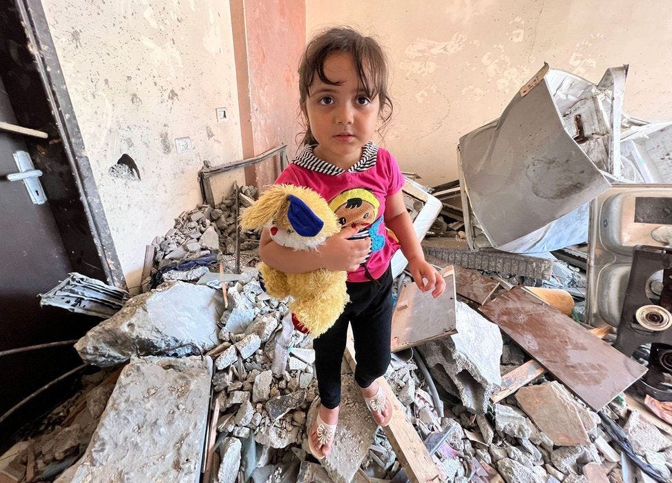 A beautiful three-year old little girl standing on rubble with a stuffed animal looks into the camera.
