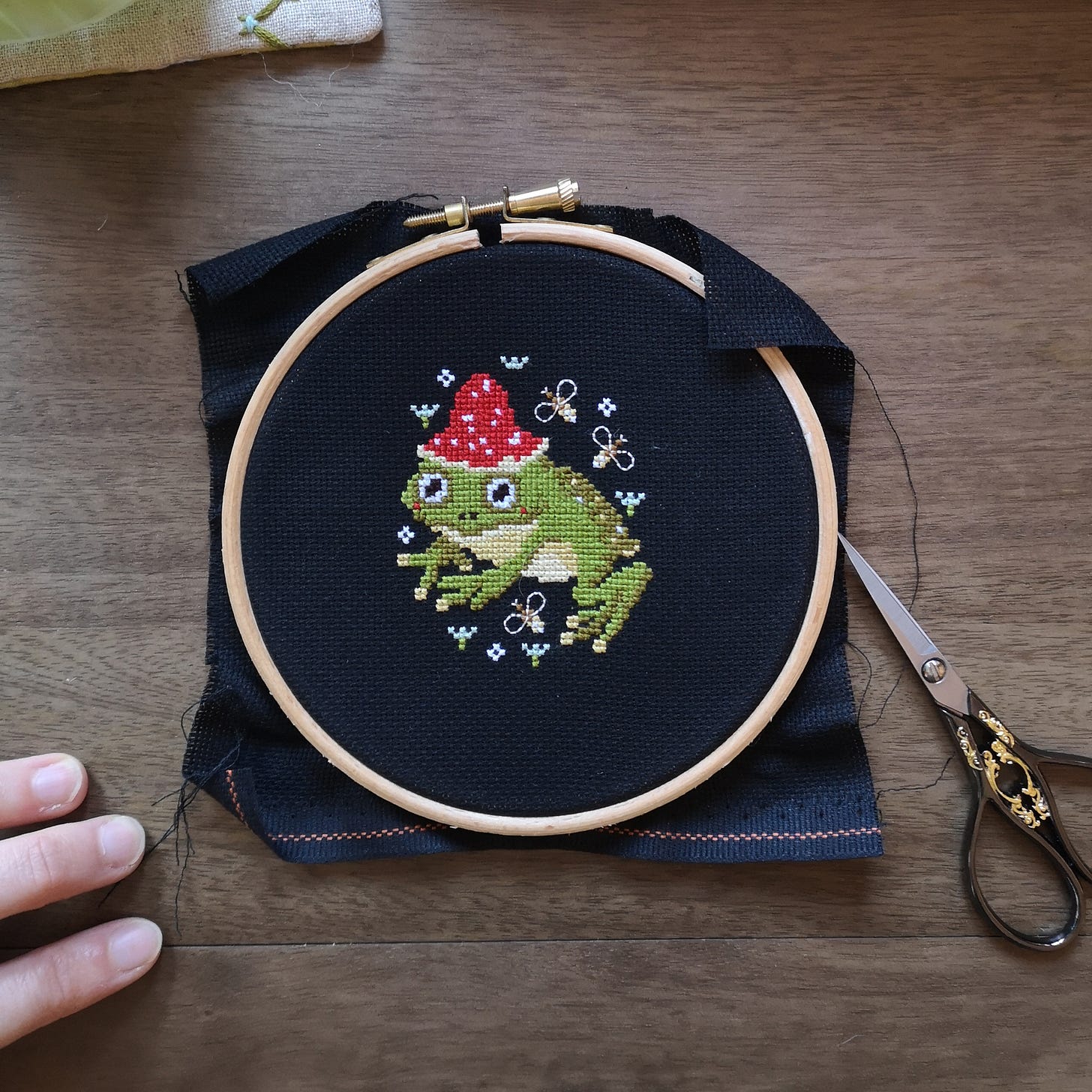 A small embroidery hoop containing black aida fabric and a cross stitched green frog wearing a red spotted mushroom for a cap. The frog is surrounded by bees and flowers. 