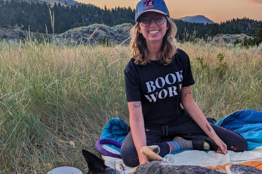 A young blonde woman wearing a baseball cap sits in tall grass on a blanket at sunset with mountains in the background and a dog rolling over for belly rubs in the foreground