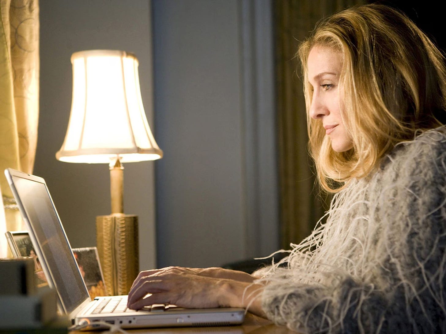 Carrie Bradshaw (Sarah Jessica Parker) sits at a desk typing on a laptop.