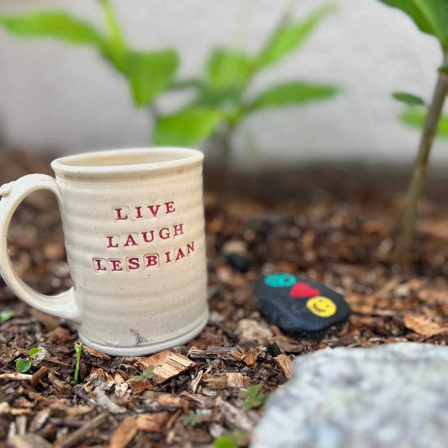 A tan ceramic mug that says "Live, Laugh, Lesbian" in red letters, sitting on ground covered in mulch with green plants in the background and a black stone with a heart, peace sign, and smiley face on it.