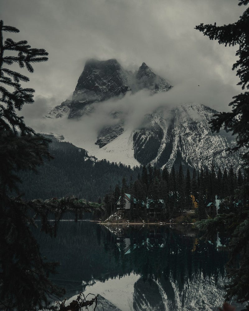 A picture of a mountain with clouds round its peak, towering above a lake, with pine trees about it.