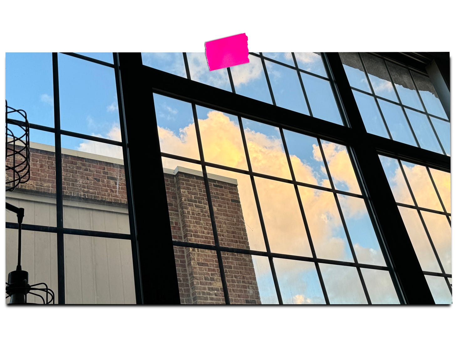 a view looking out large industrial windows at a brick wall and beyond that a blue sky with glowing orange clouds