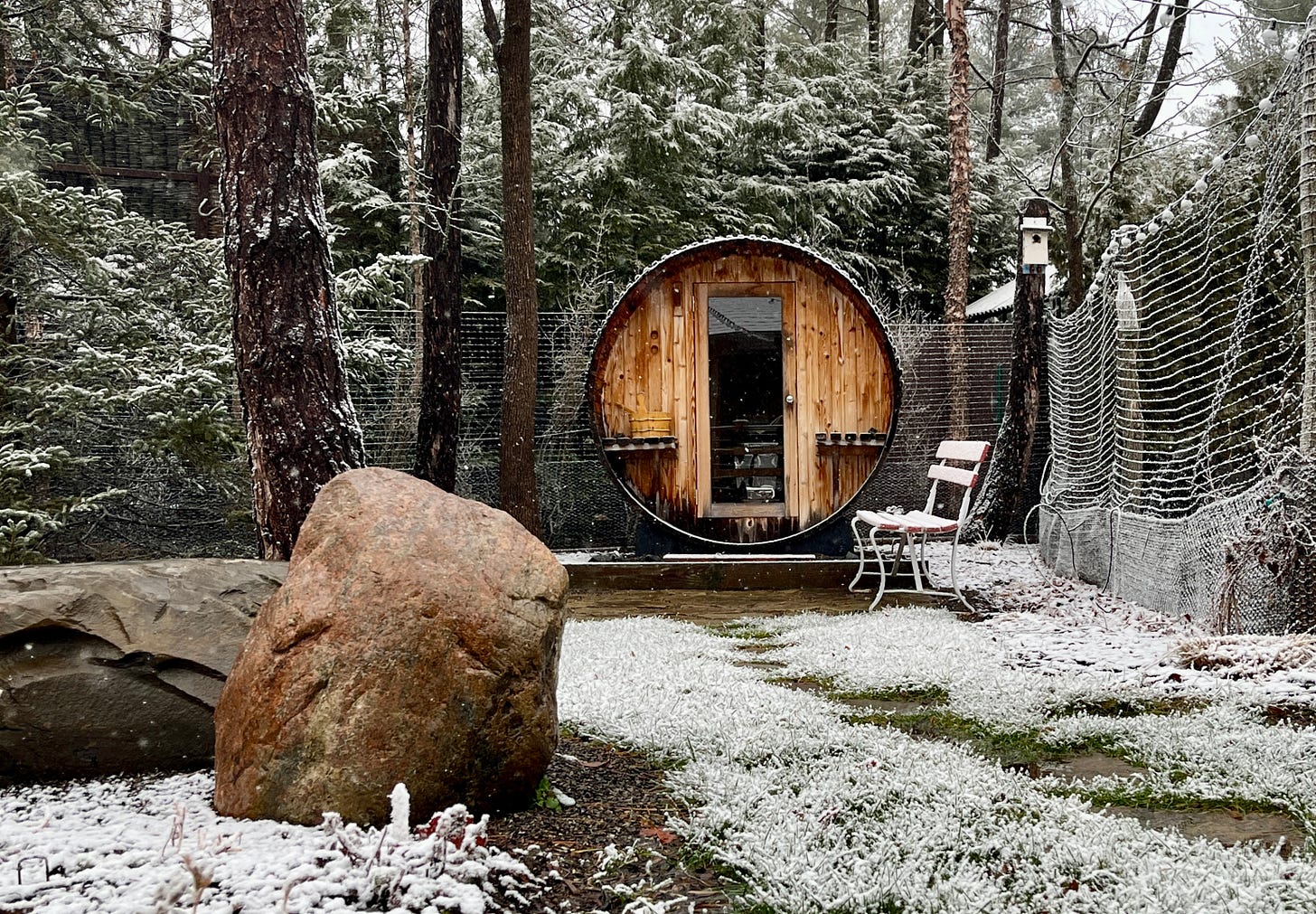 Barrel sauna with a large boulder in the foreground and a light dusting of snow on grass.