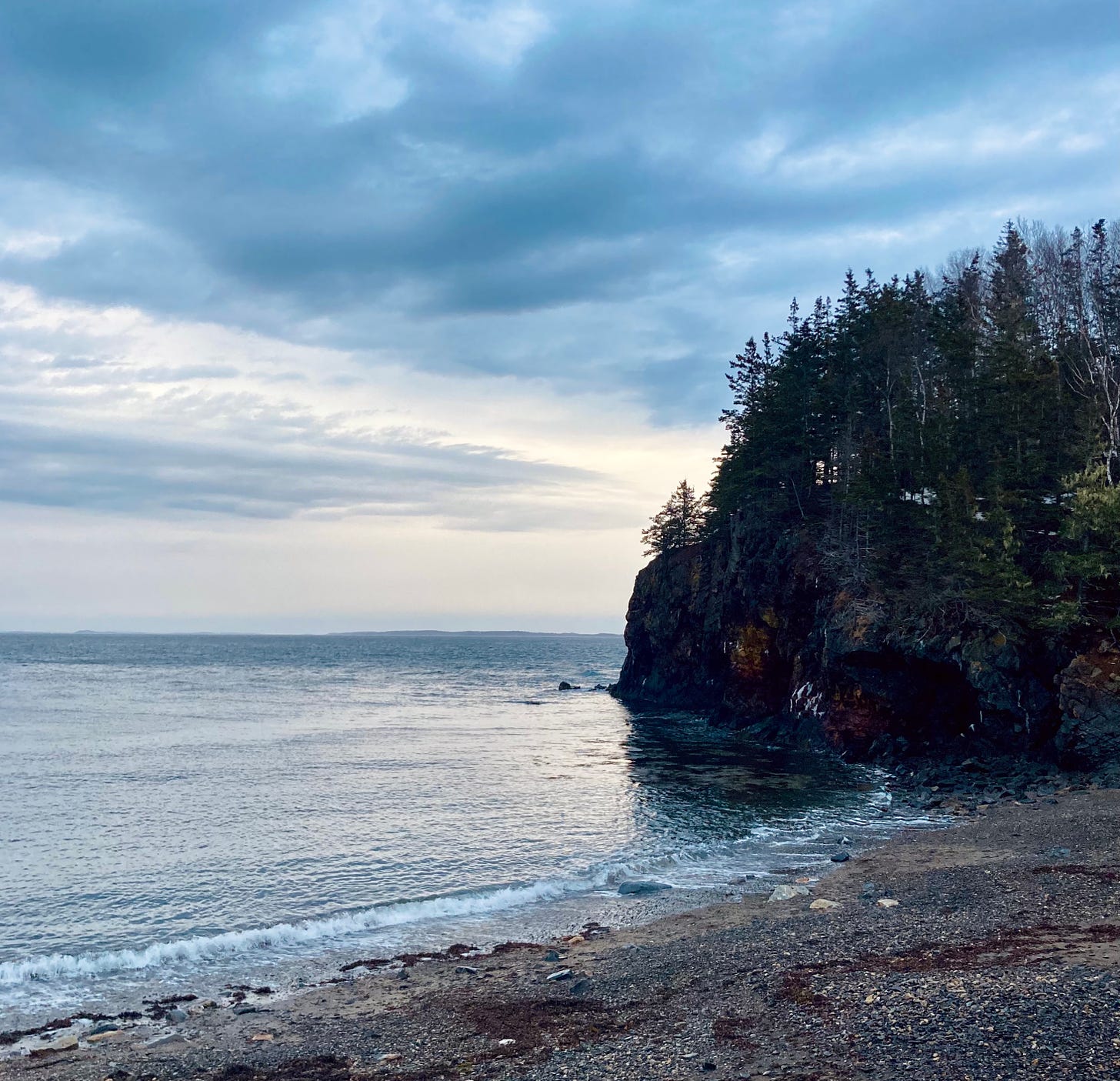 A rocky beach at sunset. The sky is overcast and illuminated. A cliff covered in evergreens juts out into the ocean from the right.