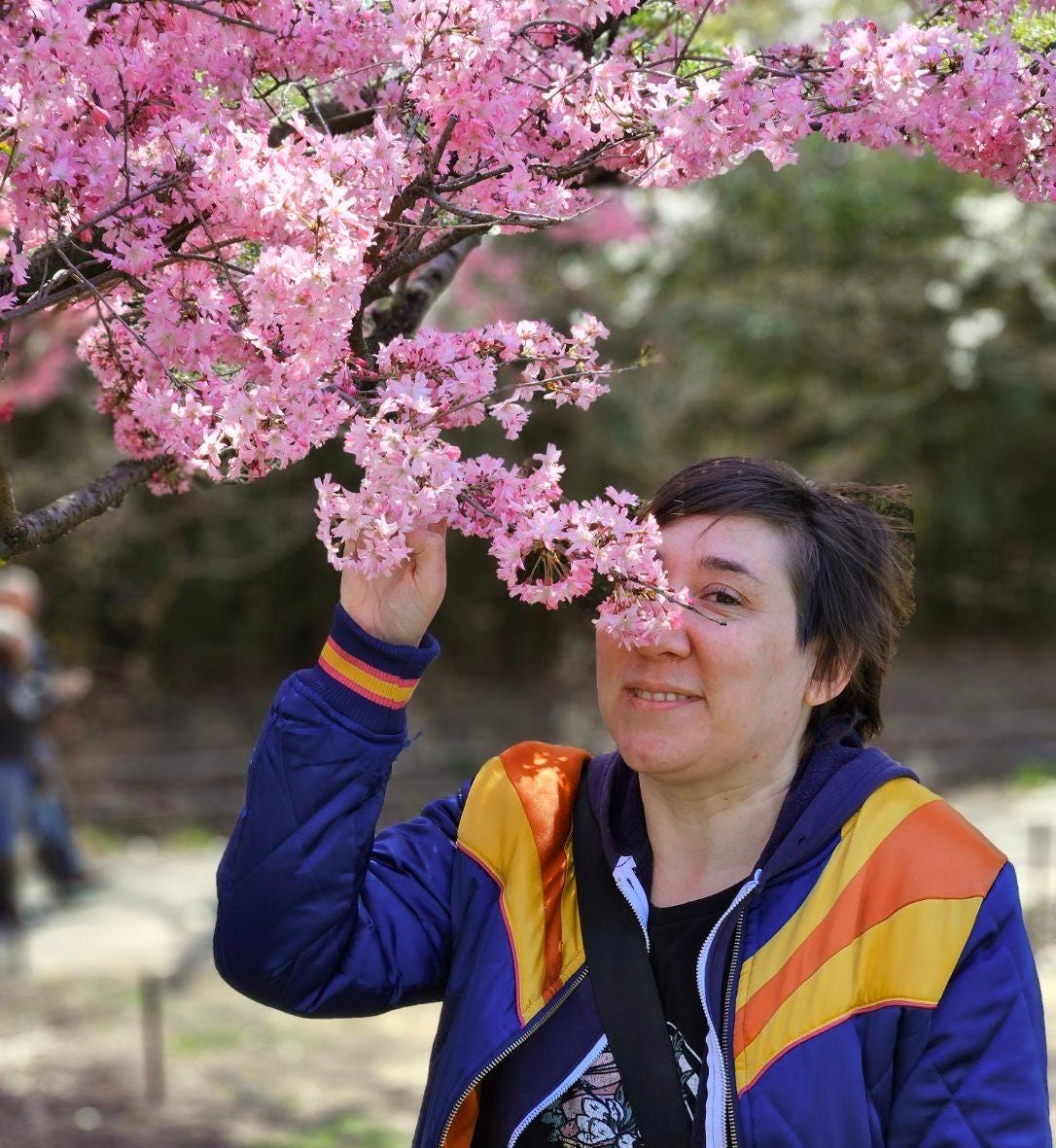 A person with short dark hair and a bright blue, yellow and orange jacket smelling a bunch of very pink cherry blossoms