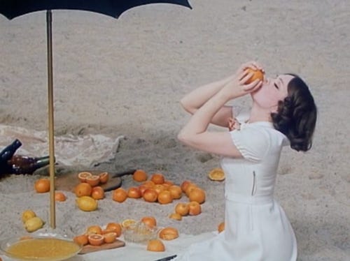 A woman in a white dress eats oranges on the beach under a black umbrella.