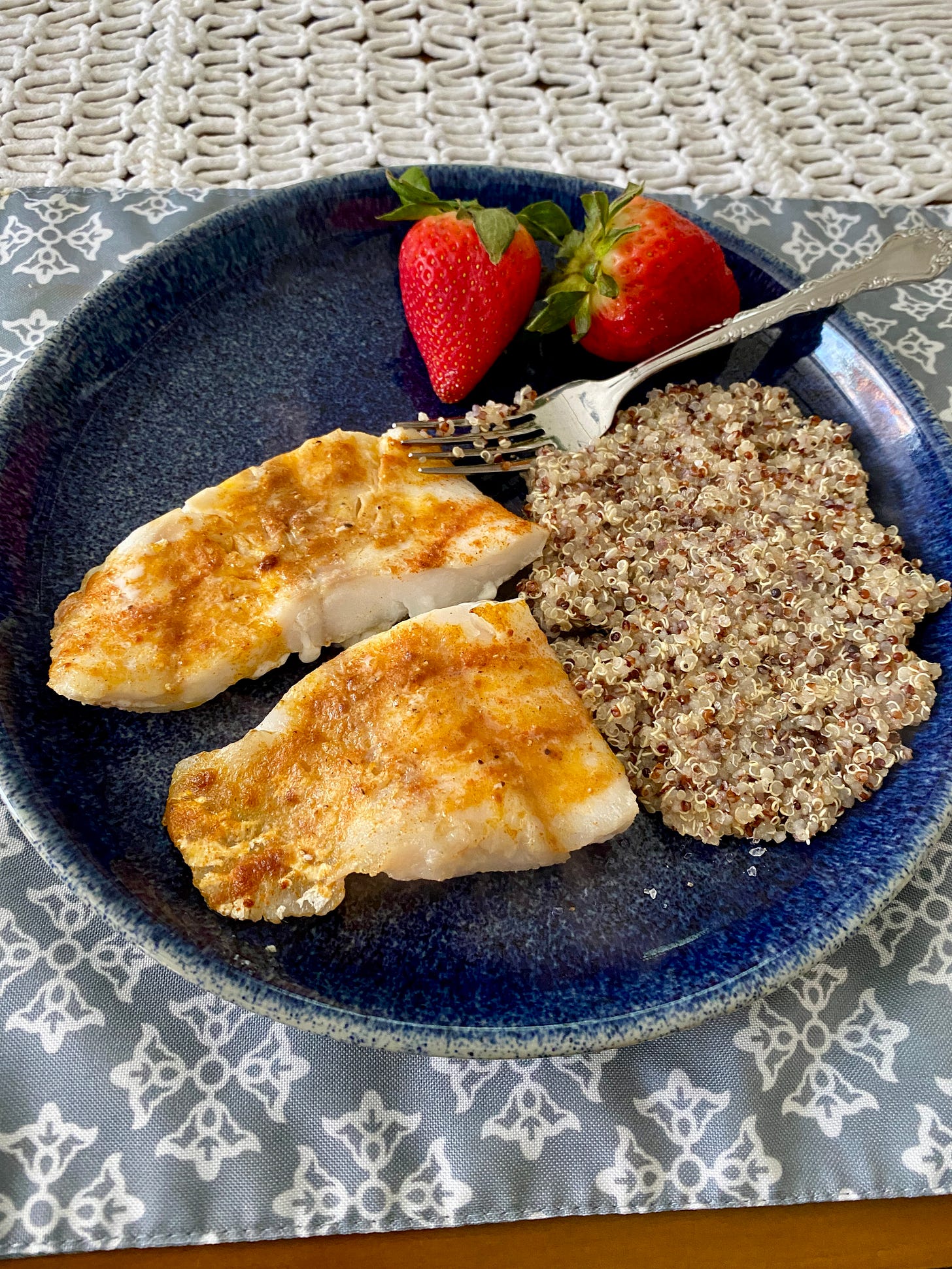 A photo of a dark blue plate sitting on a wooden table with a light blue and white placemat under it. On the plate are two pieces of cod fish, a pile of quinoa, and two strawberries.