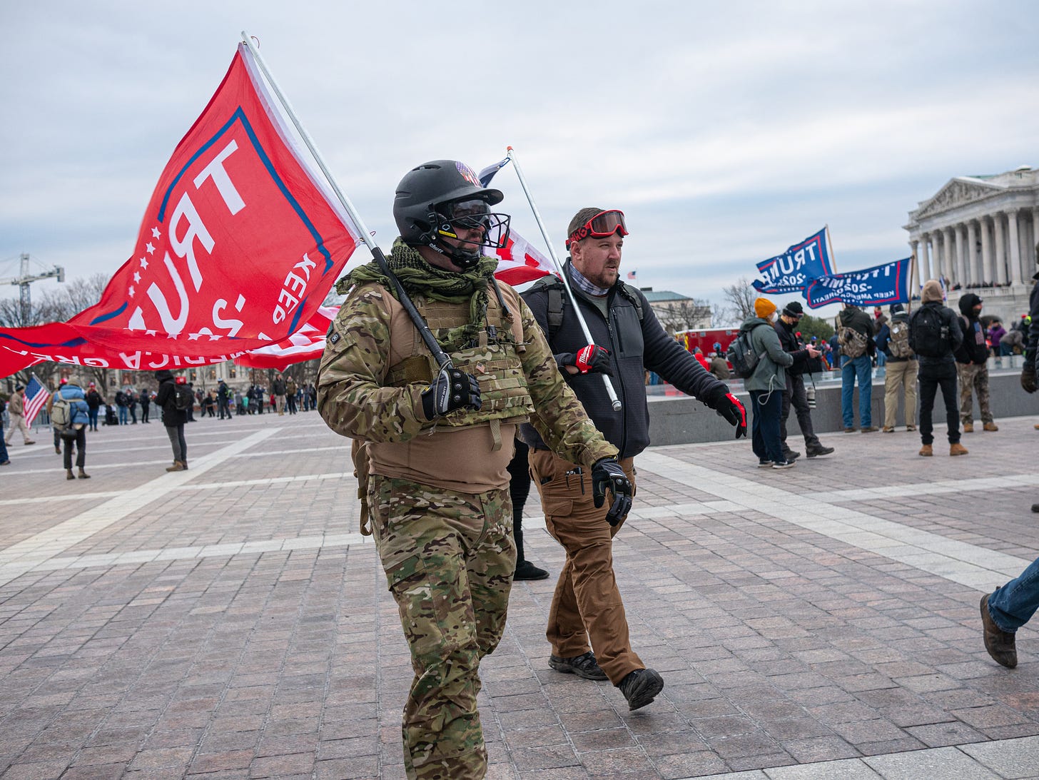 A pair of men stride with purpose on the mall in front of the US Capitol, Jan 6, 2021. One is wearing military fatigues, gloves, and what appears to be a black lacross helmet (with dark-tinted eye shield and black wire mouthguard). Over his right shoulder he is carrying a red Trump flag on a short pole. Alongside him, and in lockstep is another man in civilian clothes with a US flag on a short pole over his right shoulder. A pair or red ski-style goggles rests on his forehead. In the background, others mill about.