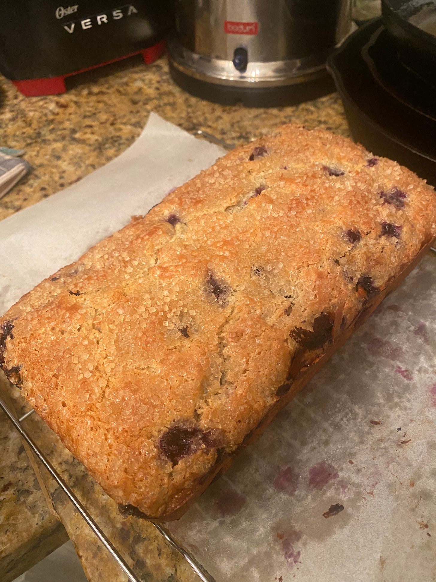 On a piece of parchment on a cooling rack, a blueberry muffin loaf, golden in colour with cooked blueberries visible in some places, The top is coated in coarse turbinado sugar.