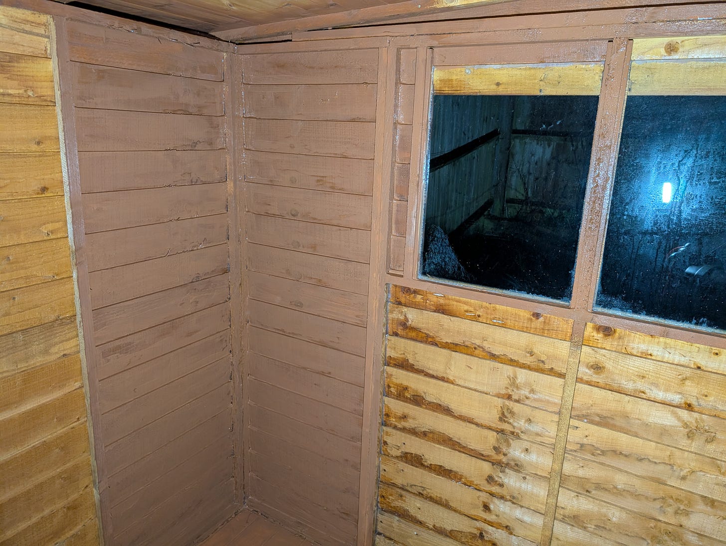 A shed corner being painted, showing patchy, slow progress with a brush.
