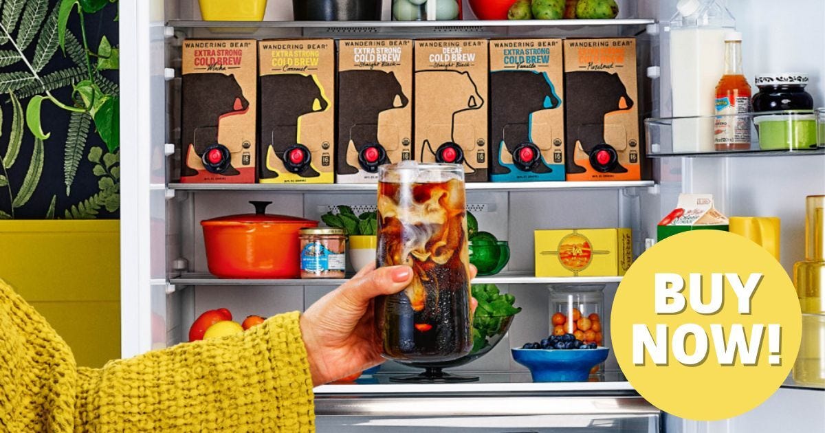 A glass of cold brew is held in front of a fridge full of on-tap Wandering Bear cold brew boxes.