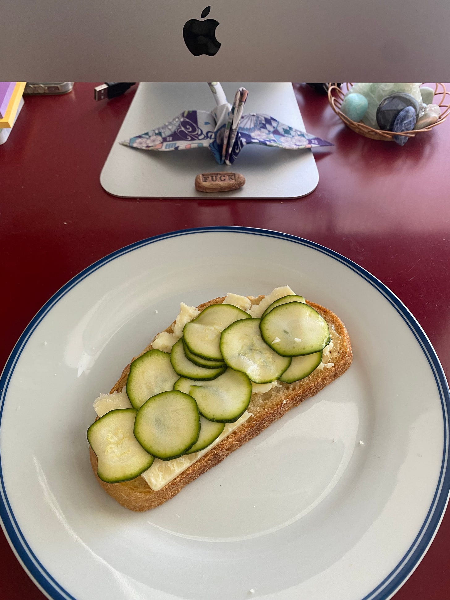 On my desk in front of my computer, a white plate with a blue rim holding an open-faced cheese and zucchini pickle sandwich on sourdough.