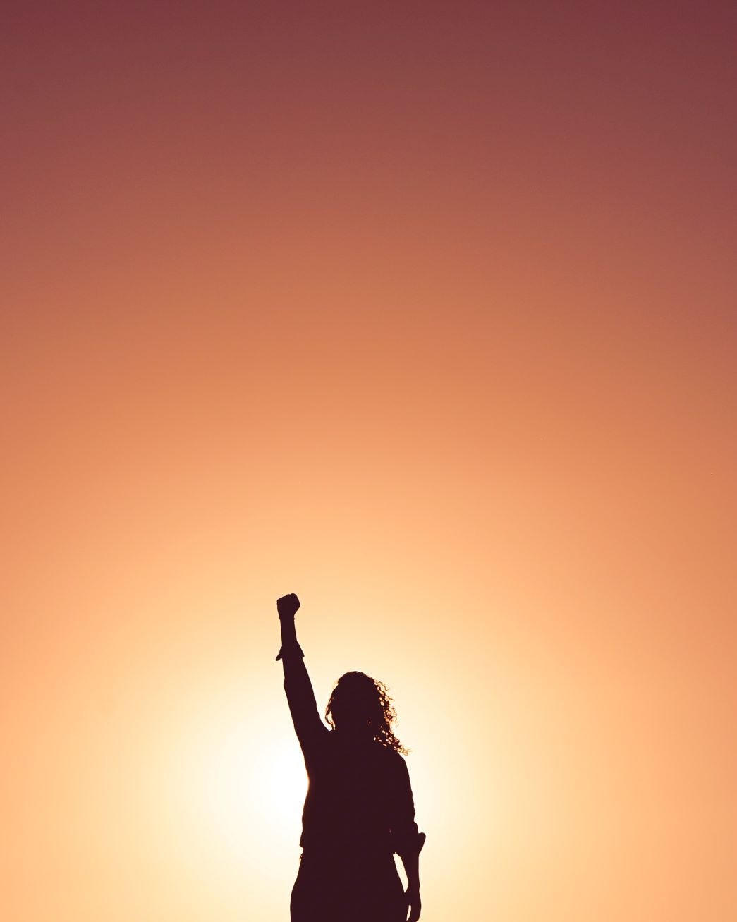 A woman is silhouetted against an orange sky. She stands alone, one arm raised above her head, her hand clenched into a fist. 