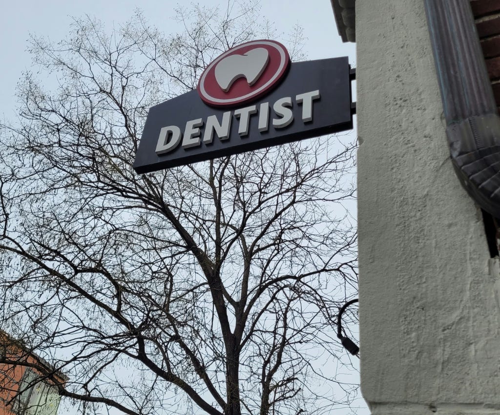 Wooden dark grey sign shaped like a section of mouth gum sticking out from a concrete wall with a brown gutter going up it.  On the sign reads “dentist” in all white caps, with a red circle at the top of the sign, with a white molar inside in circle. A large leafless tree stands against a grey-blue sky behind the DENTIST sign on the city street.