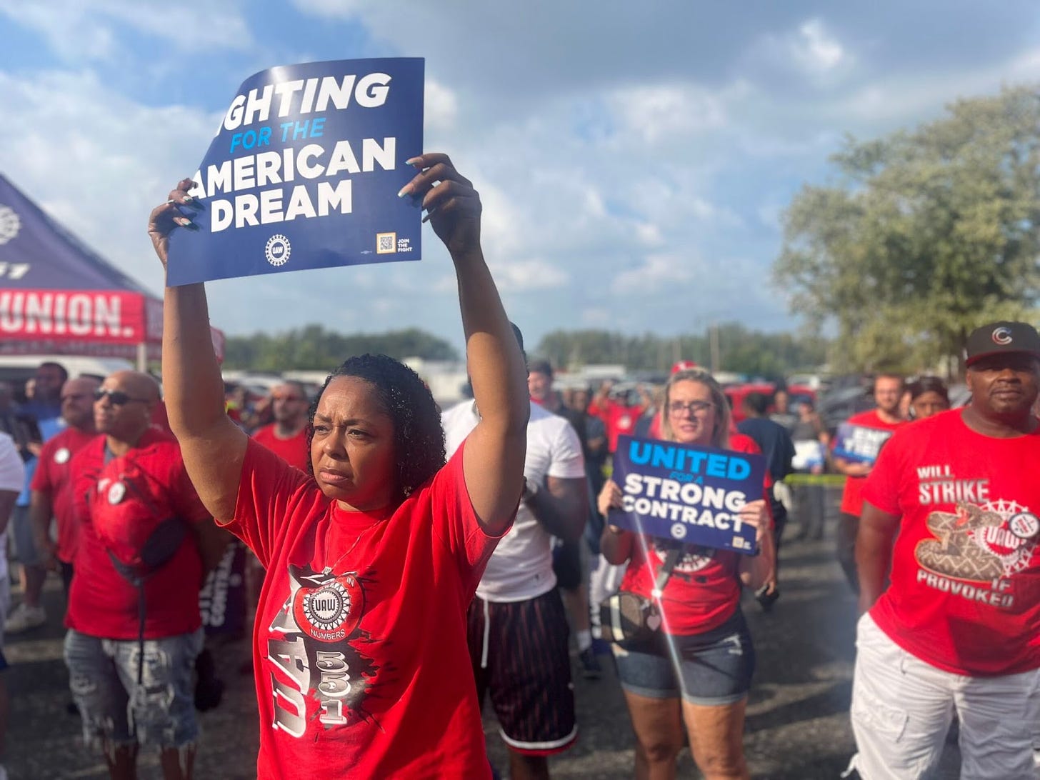 A Black woman wearing a red shirt stands in front of other people wearing red UAW shirts holding blue posters reading "Fighting for the American dream"