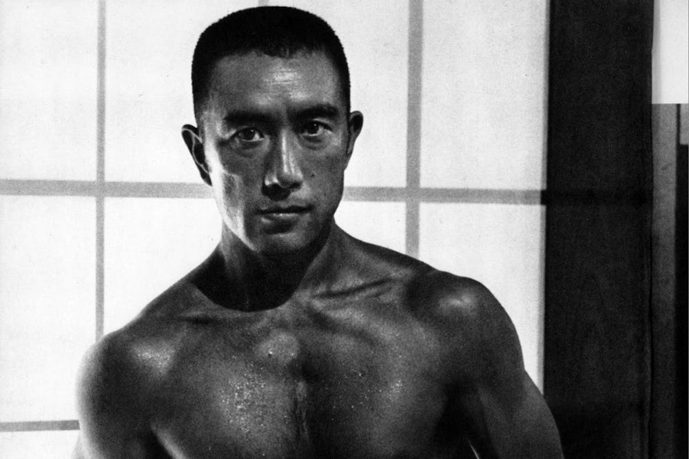 Uma foto em preto e branco de Yukio Mishima sem camisa olhando para a câmera com uma expressão séria.