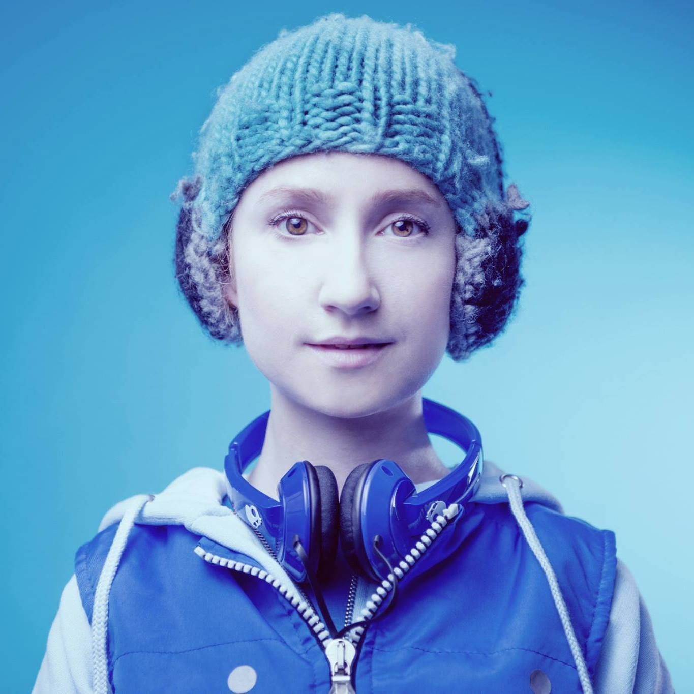 woman dressed as a 15 year old in a woollen hat with knitted in headphones and headphones around her neck. Blue tone to the whole image