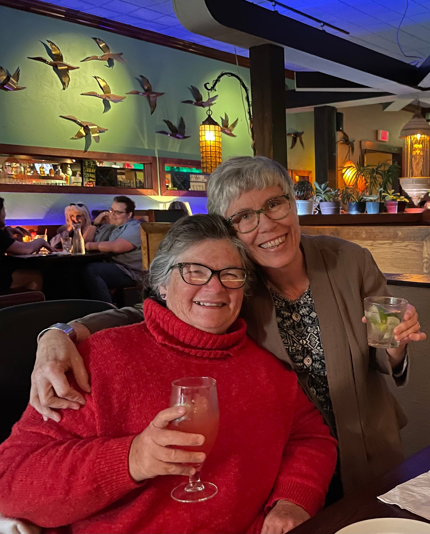 Two white woman in a restaurant smiling and hugging. One is seated in a wheelchair and the other is standing. Both have drinks in their hands. 