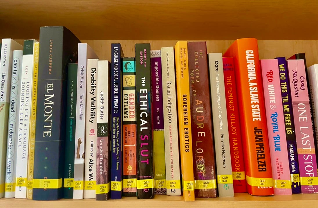 Photograph of a bookshelf. The case is made of light brown wood, and the books line up across the frame. Each one is a different color (white, yellow, black, green, black and pink, purple, orange, etc.) All books have a pink sticker at the bottom of their spine that has an alphanumeric code to identify the book in the library.