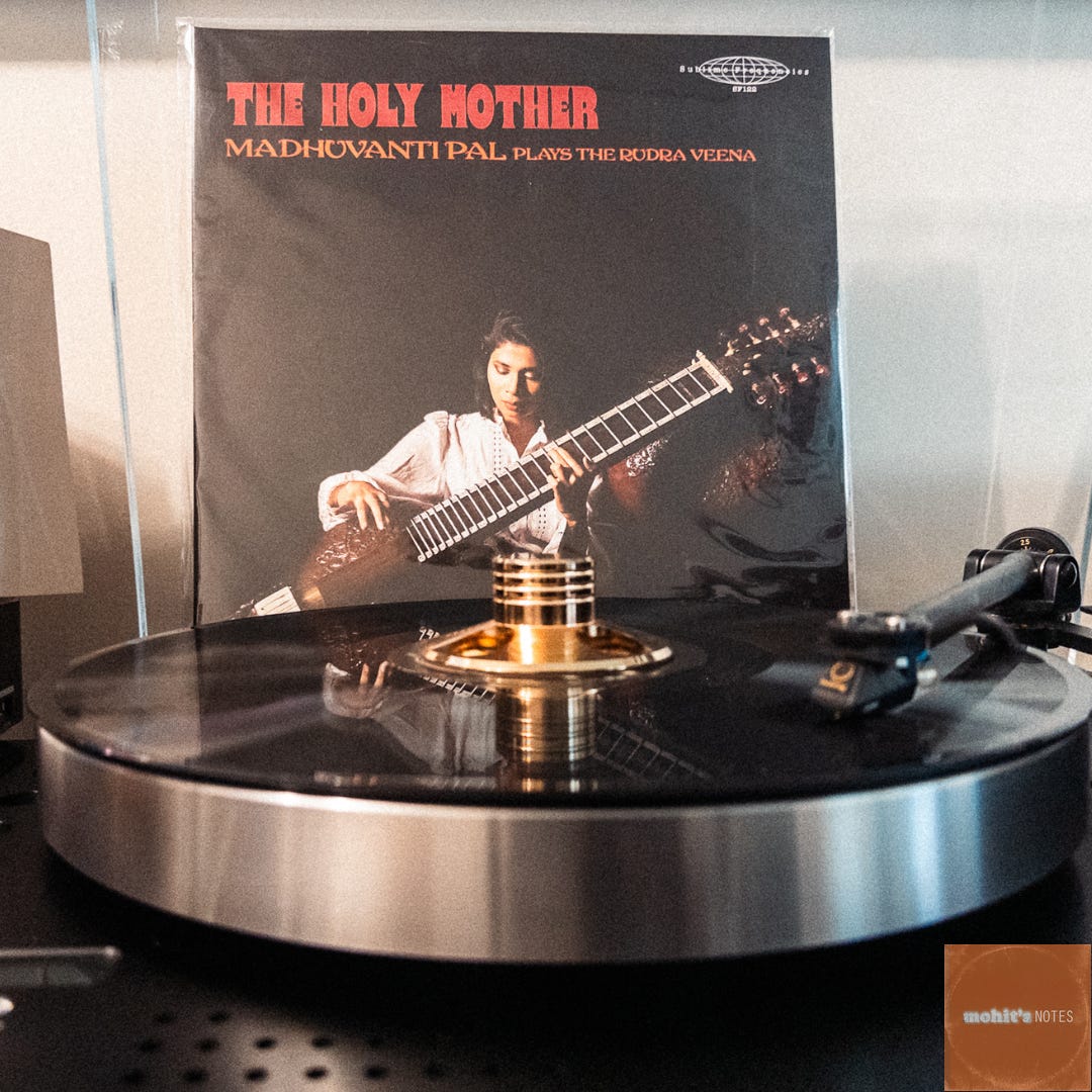 A vinyl record spins on a modern turntable, displaying the album "The Holy Mother (Plays the Rudra Veena)" by Madhuvanti Pal. The album cover features a striking black and white photograph of a musician in profile, intently focused on playing the rudra veena, a traditional Indian string instrument. The album title appears in bold red text against the dark background. The turntable's silver platter and tonearm are visible, and there appears to be a vintage-style radio partially visible in the background.