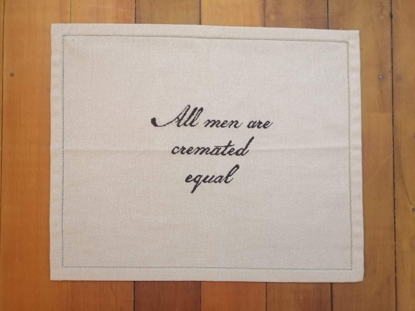 Cream linen placemat with dark brown text in a handwritten script "All men are cremated equal"