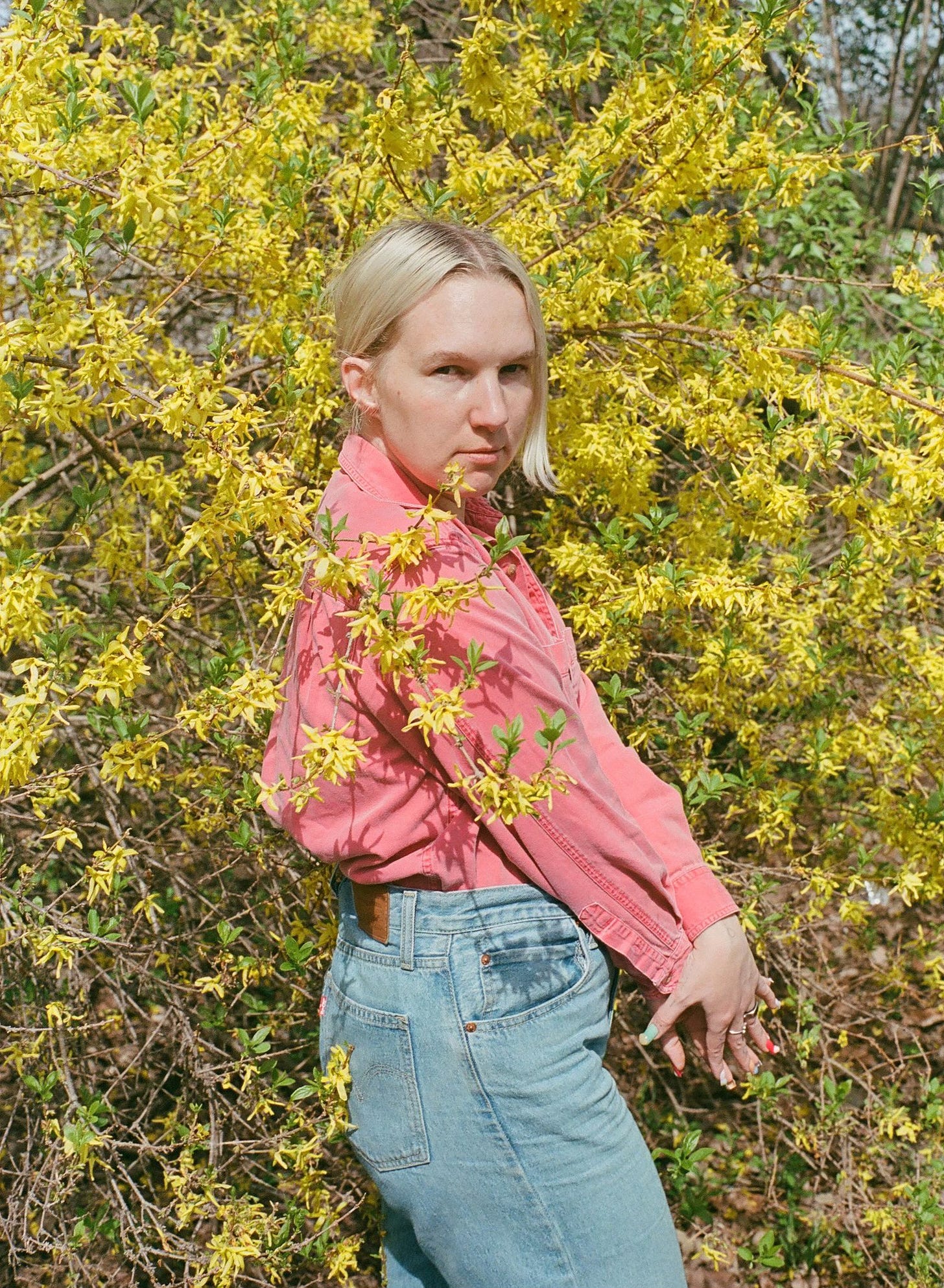 A person with short blonde hair, wearing a pink shirt and jeans, stands in front of bright yellow flowering bushes, looking directly at the camera.