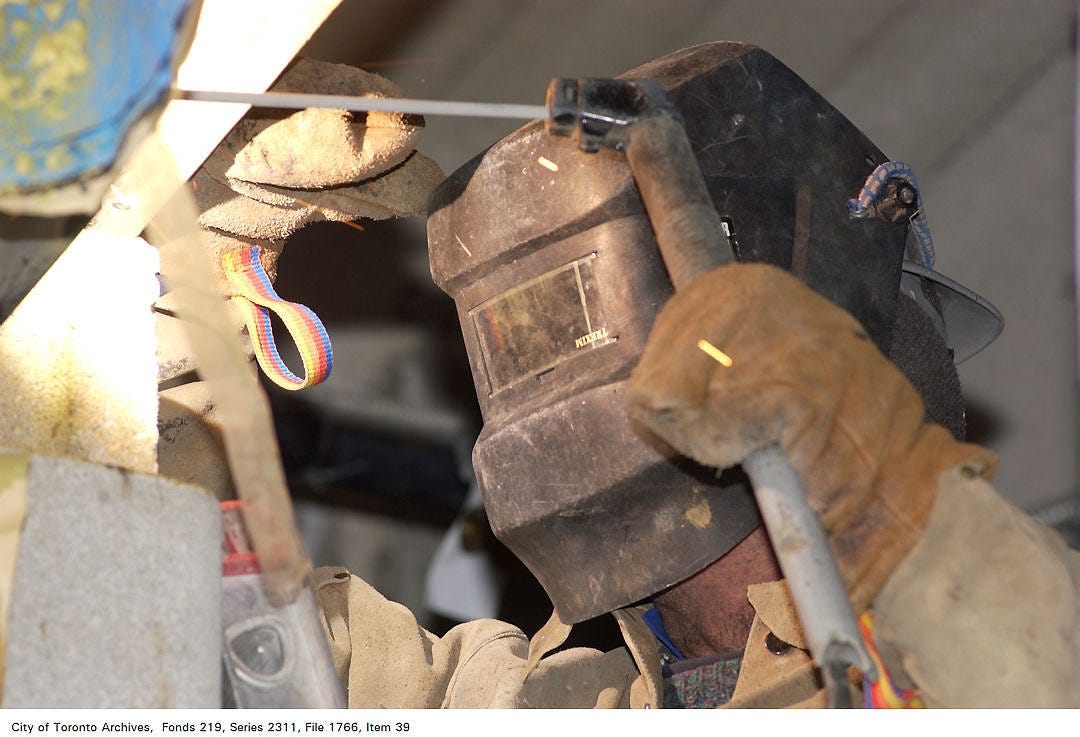 A Toronto Archives photo of a contractor doing work for the City. The worker is wearing heavy gloves and a black welder's mask, holding a tool