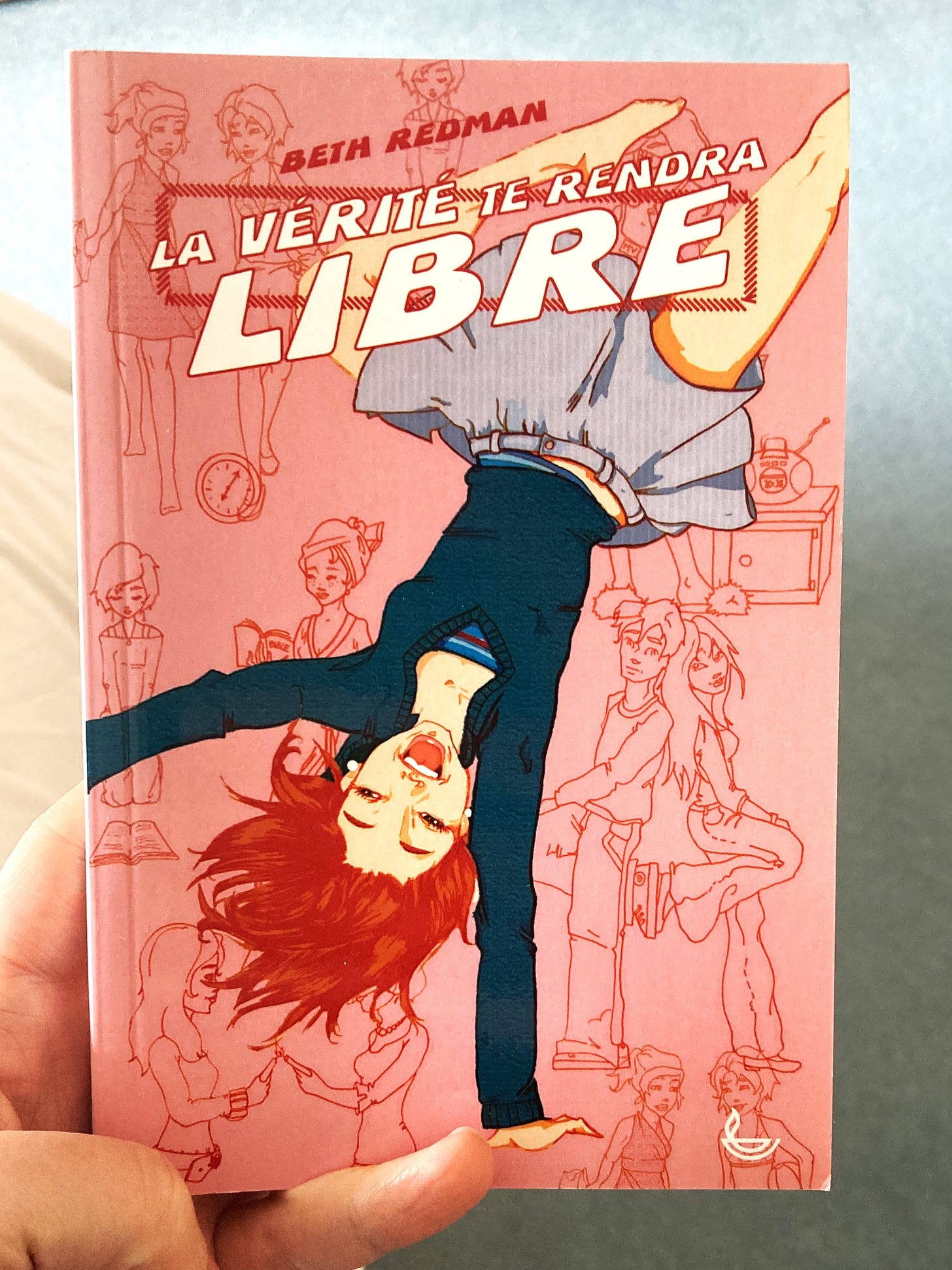 The book cover is pink. Across the front is a illustrated white girl wearing a blue jumper and skirt (somehow defying gravity as she is upside down).