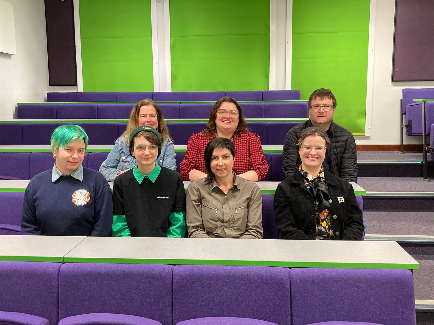 Photograph of sevenfour women, one man and two non-binary people sitting in a green and purple lecture theatre smiling at the camera. Despite it being an EDI panel, everyone is white. Yikes. 