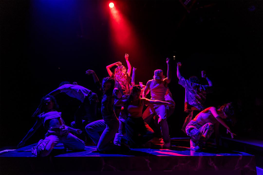 A group of actors on a darkened stage surrounding the central woman who is lit in red-purple light.