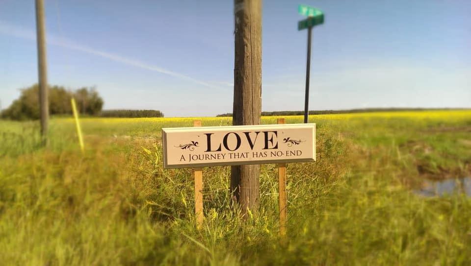 A sign that says, Love - A Journey That Has No End in a field of wheat and canola in Northern Alberta.