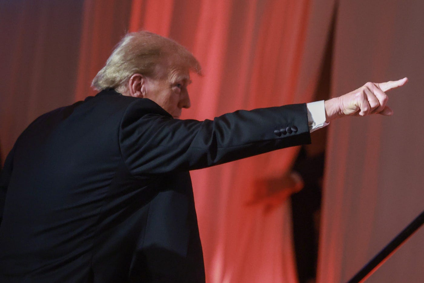 Former President Donald Trump pointing upward during a speech, with a dramatic red-lit background. The image captures his strong gesture while addressing an audience, conveying his influence in right-wing media and political discourse.