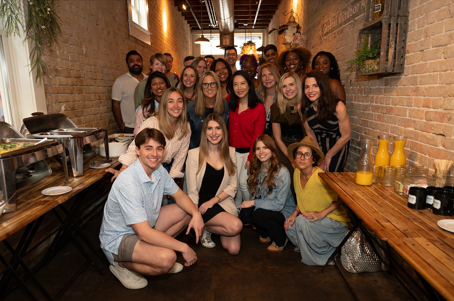 Photo of a large group of people posing and looking at the camera within a narrow restaurant space. There are brick walls and chaffing dishes on the side with breakfast tacos. The restaurant is called Waltons Fancy & Staple. The group is a mix of marketers and creators, journalists, analysts and academics 