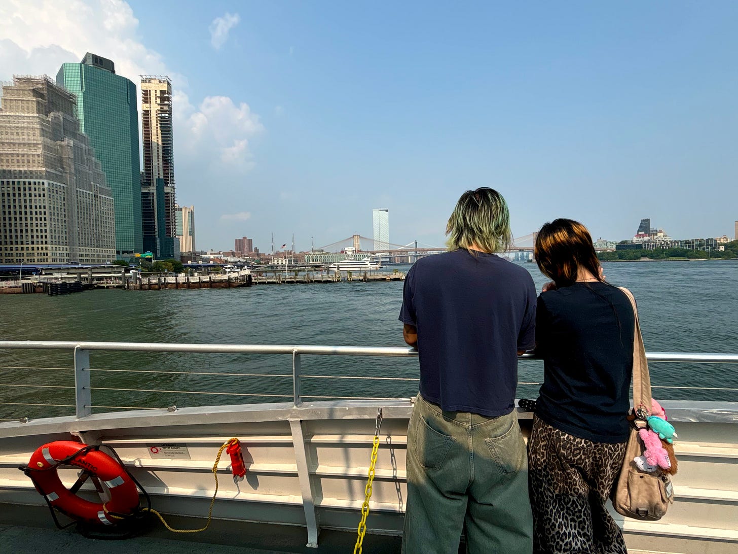 Two people standing at the railing of a ferry, watching Manhattan approach.