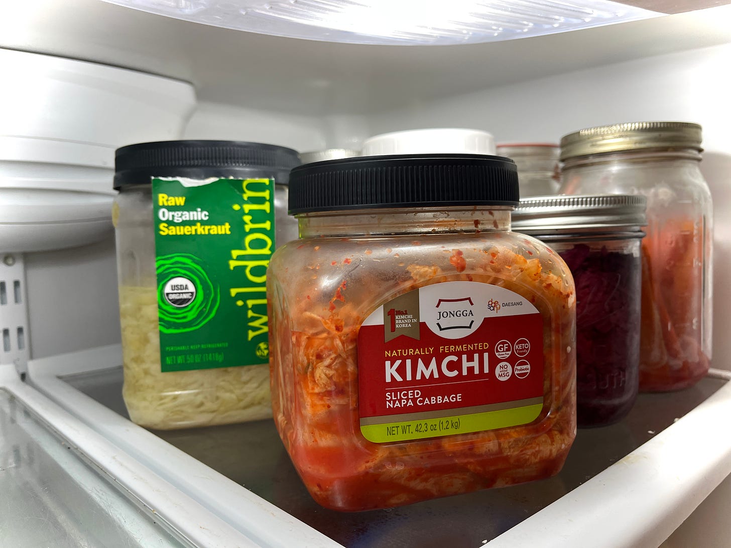 A refrigerator shelf filled with jars of fermented foods, including sauerkraut and kimchi