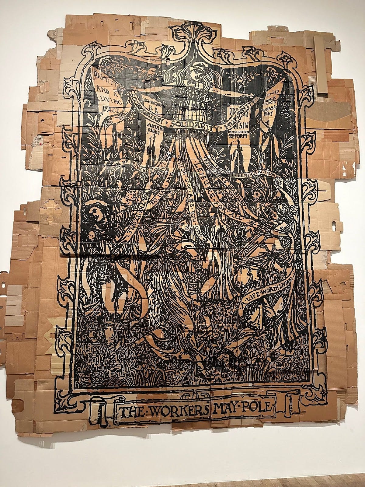 a photo of a large piece of art of a collage of cardboard with black ink design of a maypole illustration with banners reading "dignity and a living wage" "socialization solidarity humanity" and "leisure for all" "a life worth living"