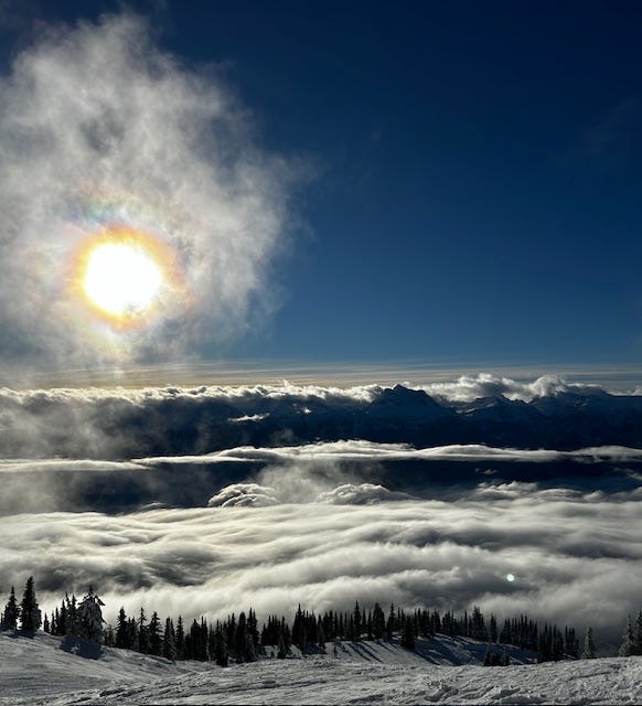 A snow-covered ski hill, trees with a bright blue sky and the sun behind wispy clouds.